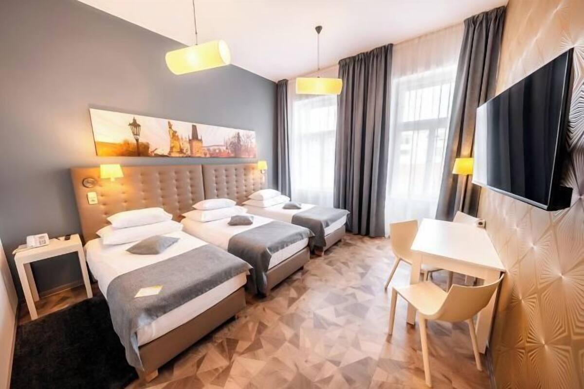 Ein Hotelzimmer mit drei Betten, einem Schreibtisch, einem Sessel, einem Flachbildfernseher und einem Bild an der Wand.