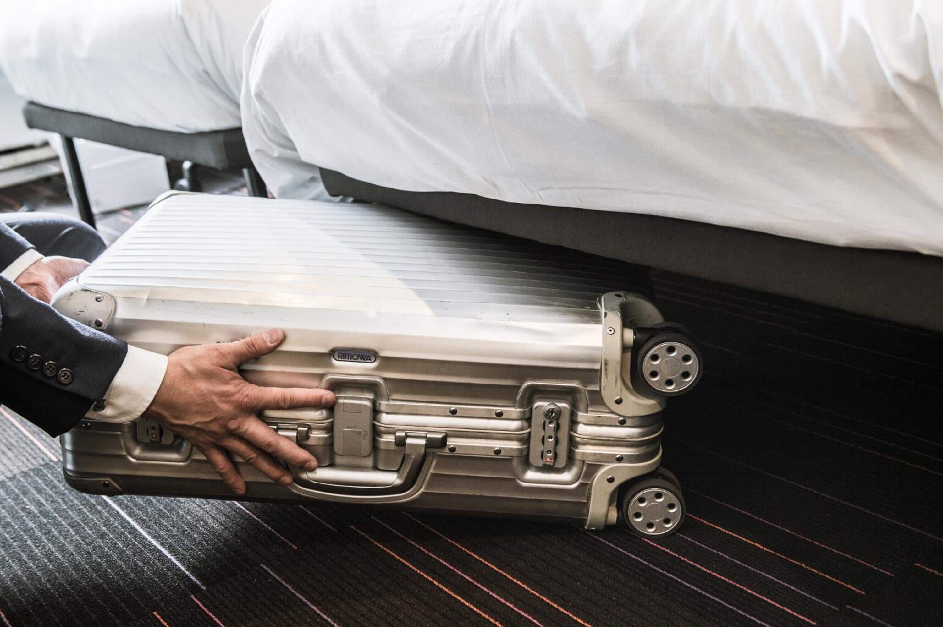 Une personne pose une valise métallique sur un lit où se trouve un oreiller blanc.