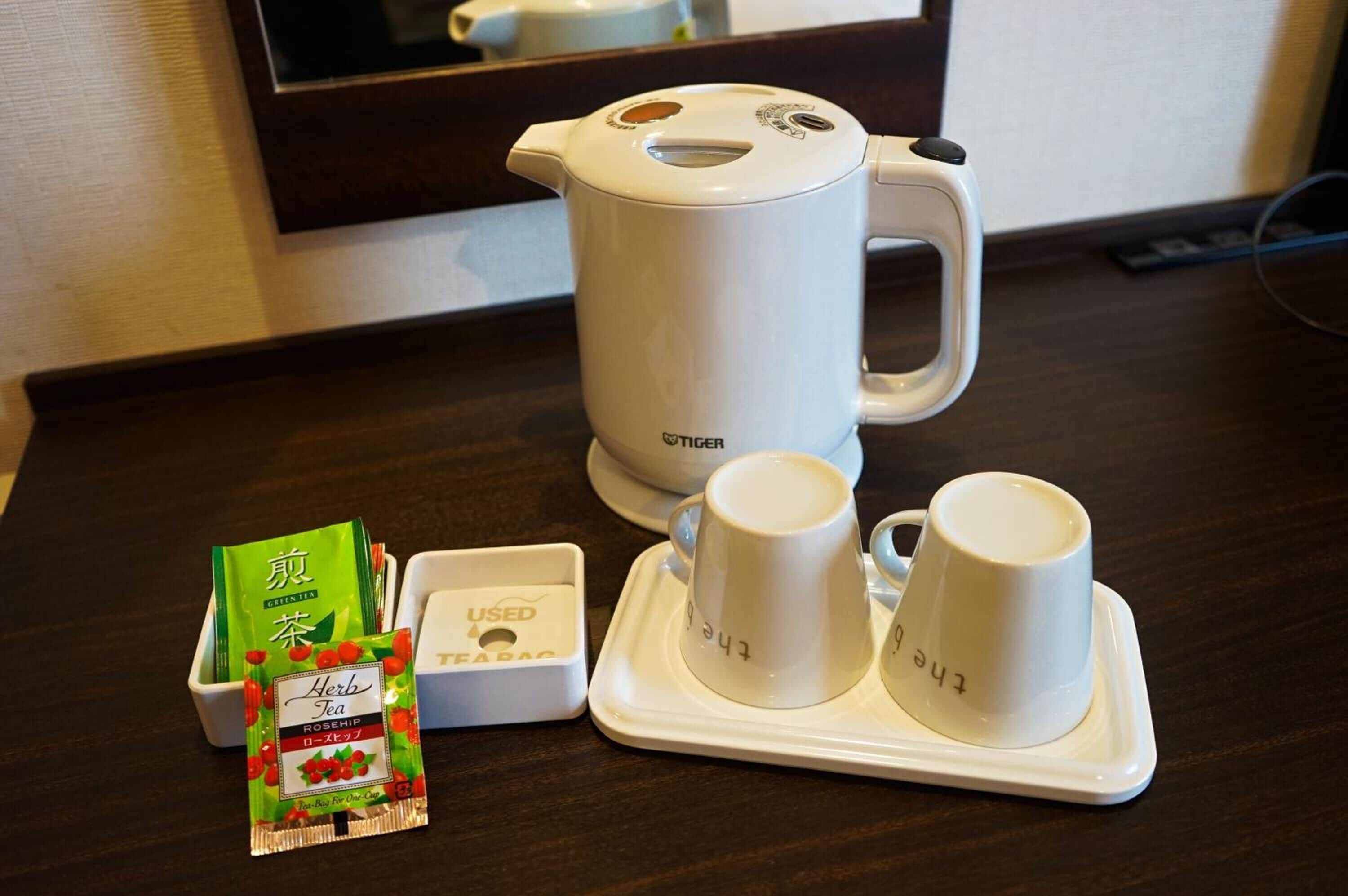 Una kitchenette con un hervidor eléctrico blanco, dos tazas blancas sobre una bandeja, un paquete de té verde y una caja con la etiqueta 'USED'.