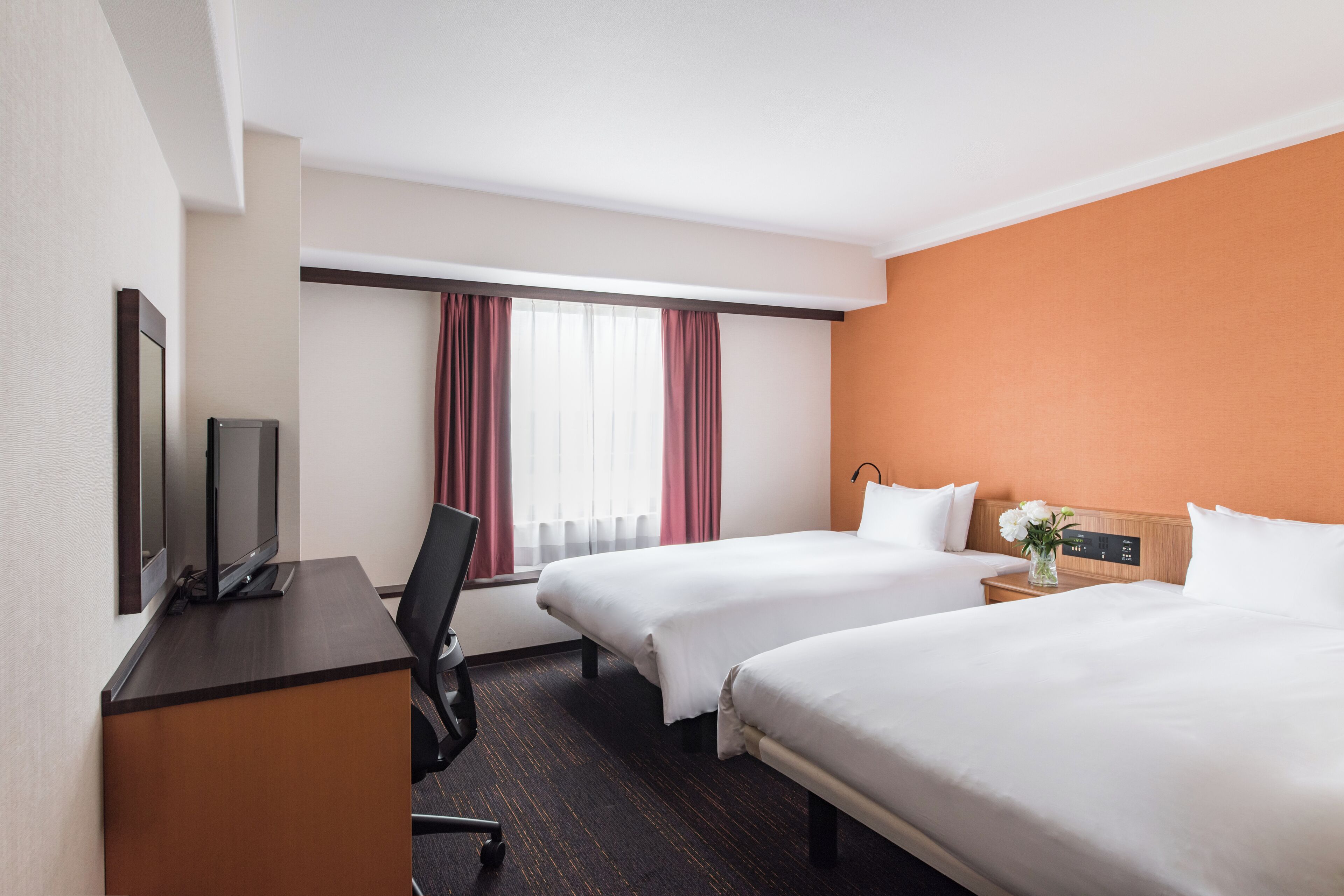 A hotel room with two beds, a desk with a TV, a chair, and a nightstand with a vase of flowers.