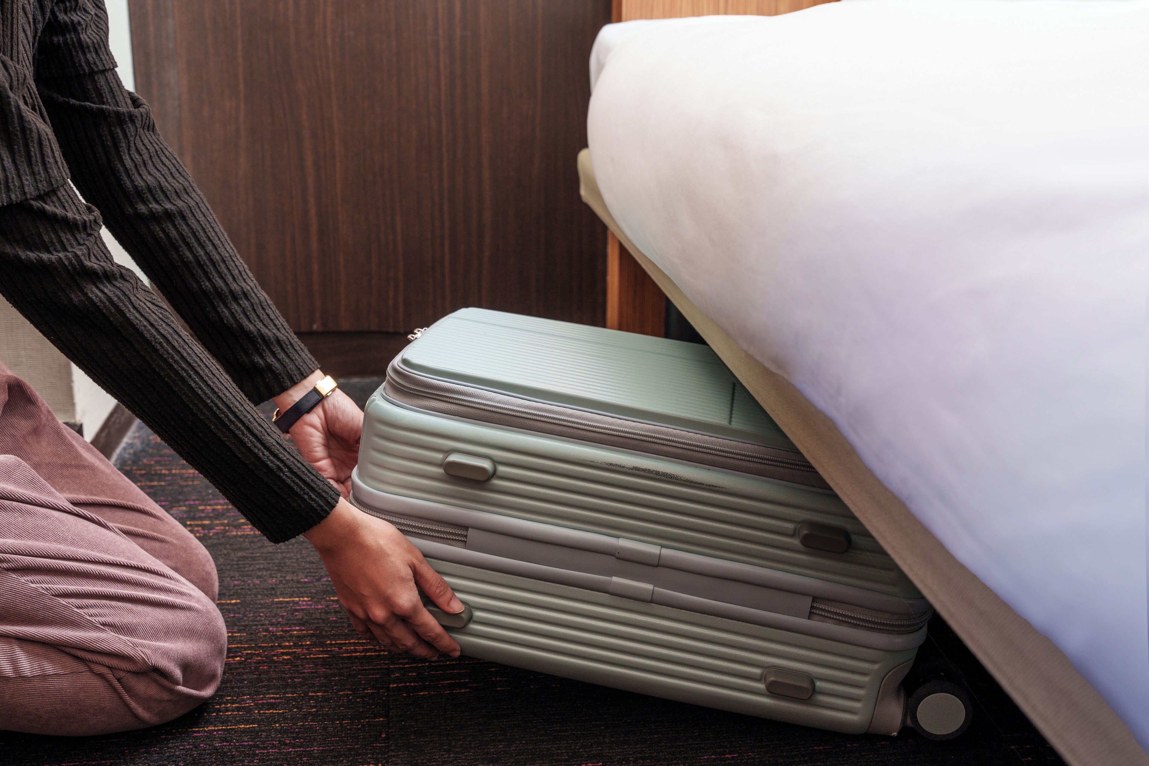 Une personne tenant une valise verte dans une pièce où se trouvent un lit et des meubles en bois.