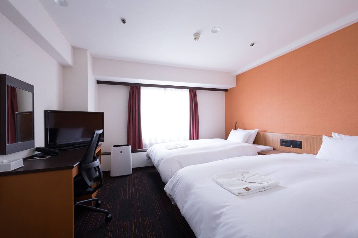 A hotel room with two beds, a desk with a computer, a TV, and a window with curtains.