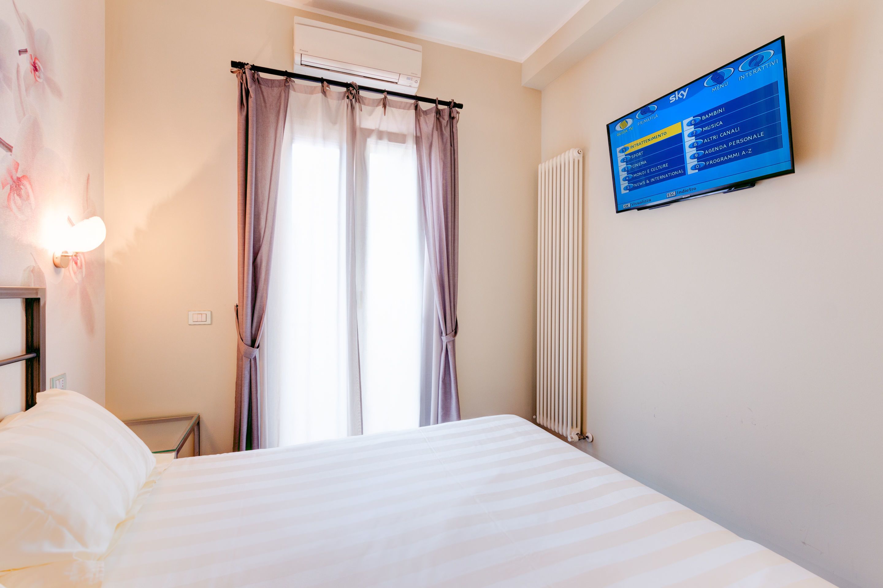 A hotel room with a bed, a wall-mounted TV displaying a menu, a window with curtains, and a radiator.