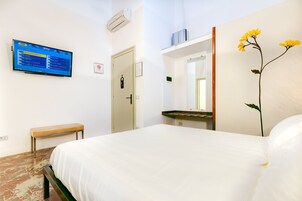 Hotelkamer met een bed, een bankje, een aan de muur bevestigde televisie waarop een programmalijst staat, een deur, een spiegel en een muur met een gele bloem.