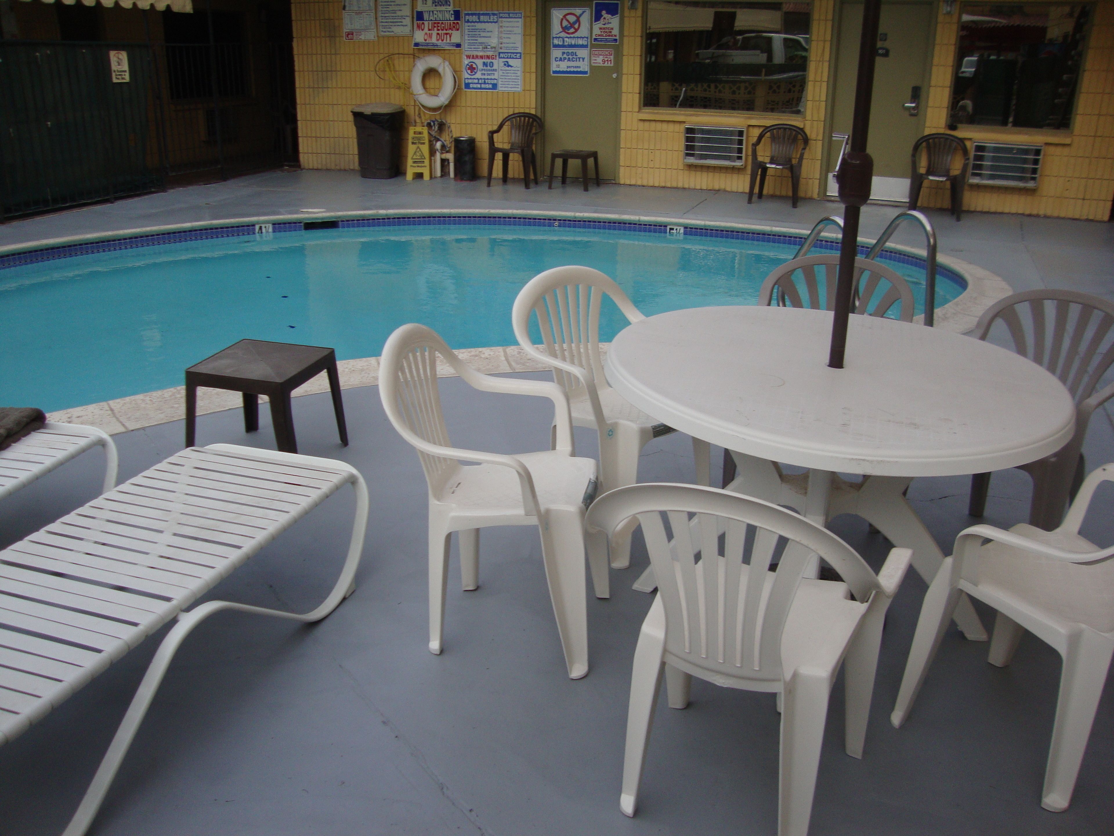 Un espace piscine intérieure avec des chaises et une table en plastique blanc, un poste de surveillance pour les sauveteurs et divers panneaux de sécurité affichés sur les murs.