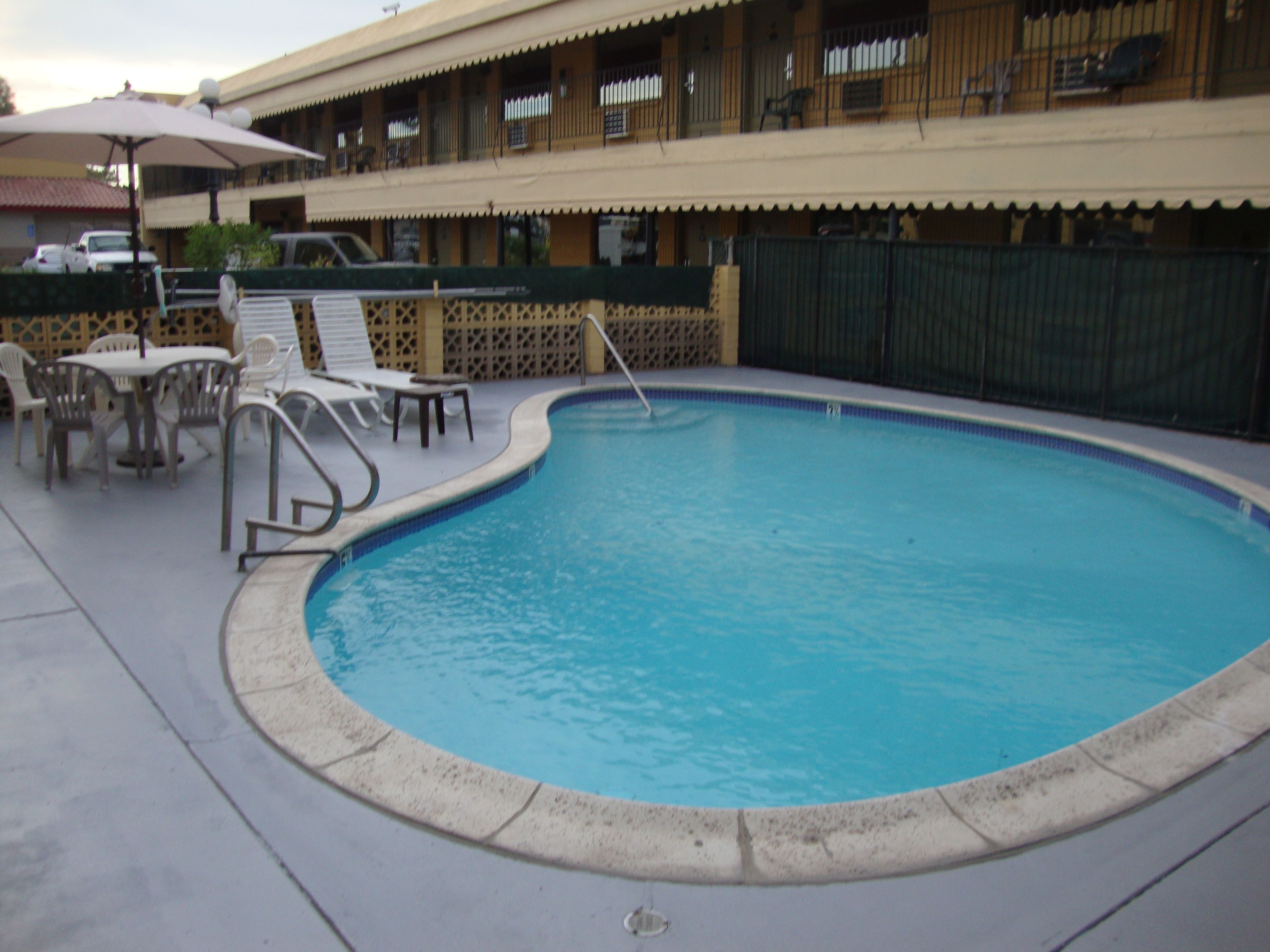A swimming pool with lounge chairs and tables, surrounded by a fence and a building with balconies.