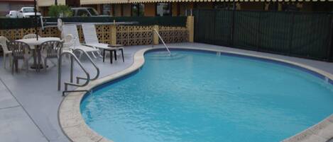 A swimming pool with lounge chairs and tables, surrounded by a fence and a building with balconies.