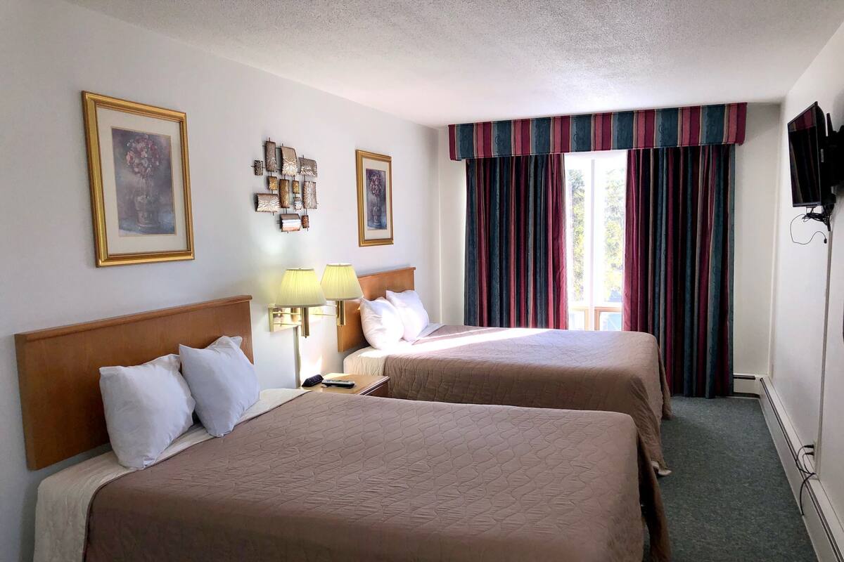 Habitación de hotel con dos camas, una mesita de noche, lámparas, televisor y ventana con cortinas.