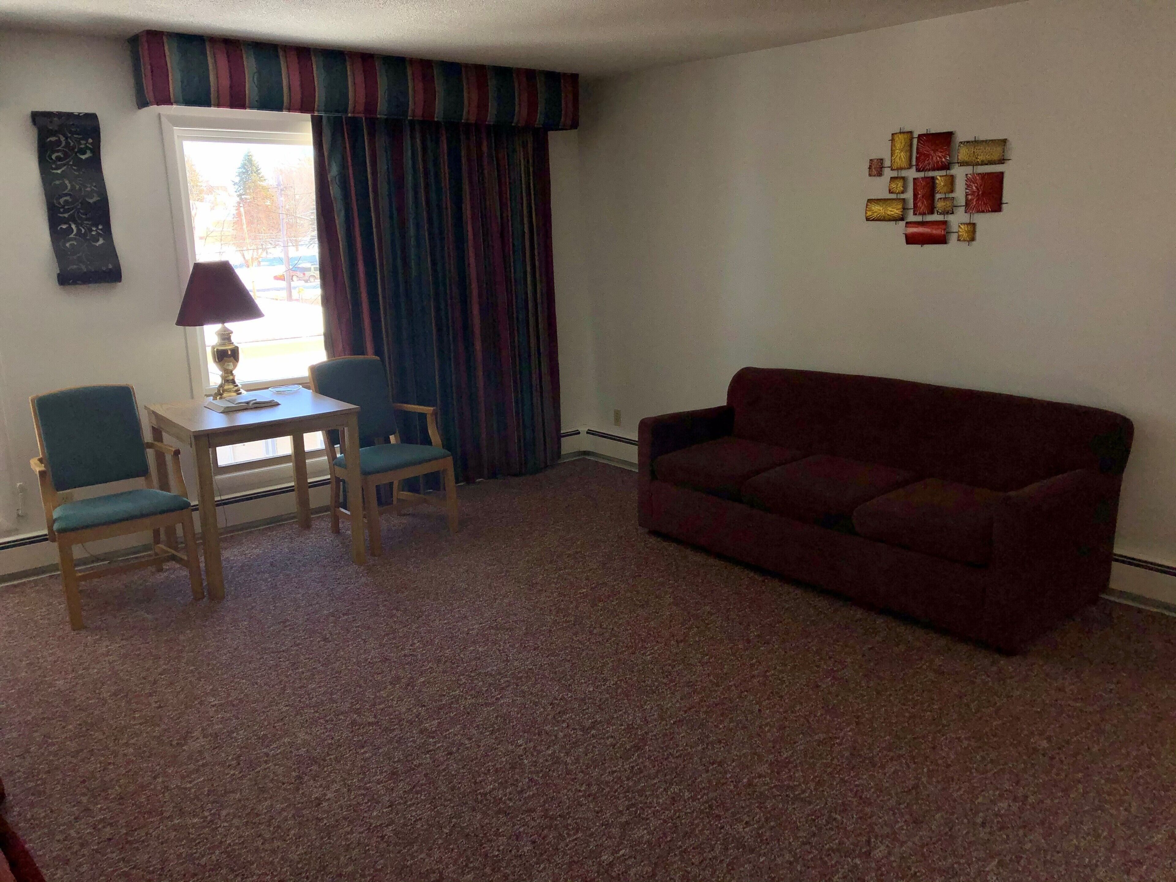 A living room with a red sofa, two chairs, a small table with a lamp, a window with curtains, and a wall decoration.