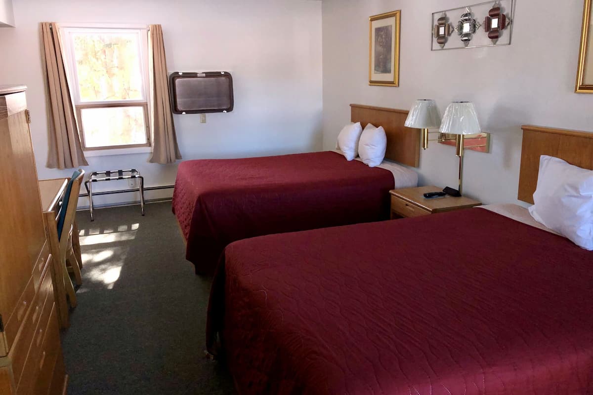 Habitación de hotel con dos camas, televisión, mesita de noche con lámpara, ventana con cortinas y un banco de madera.