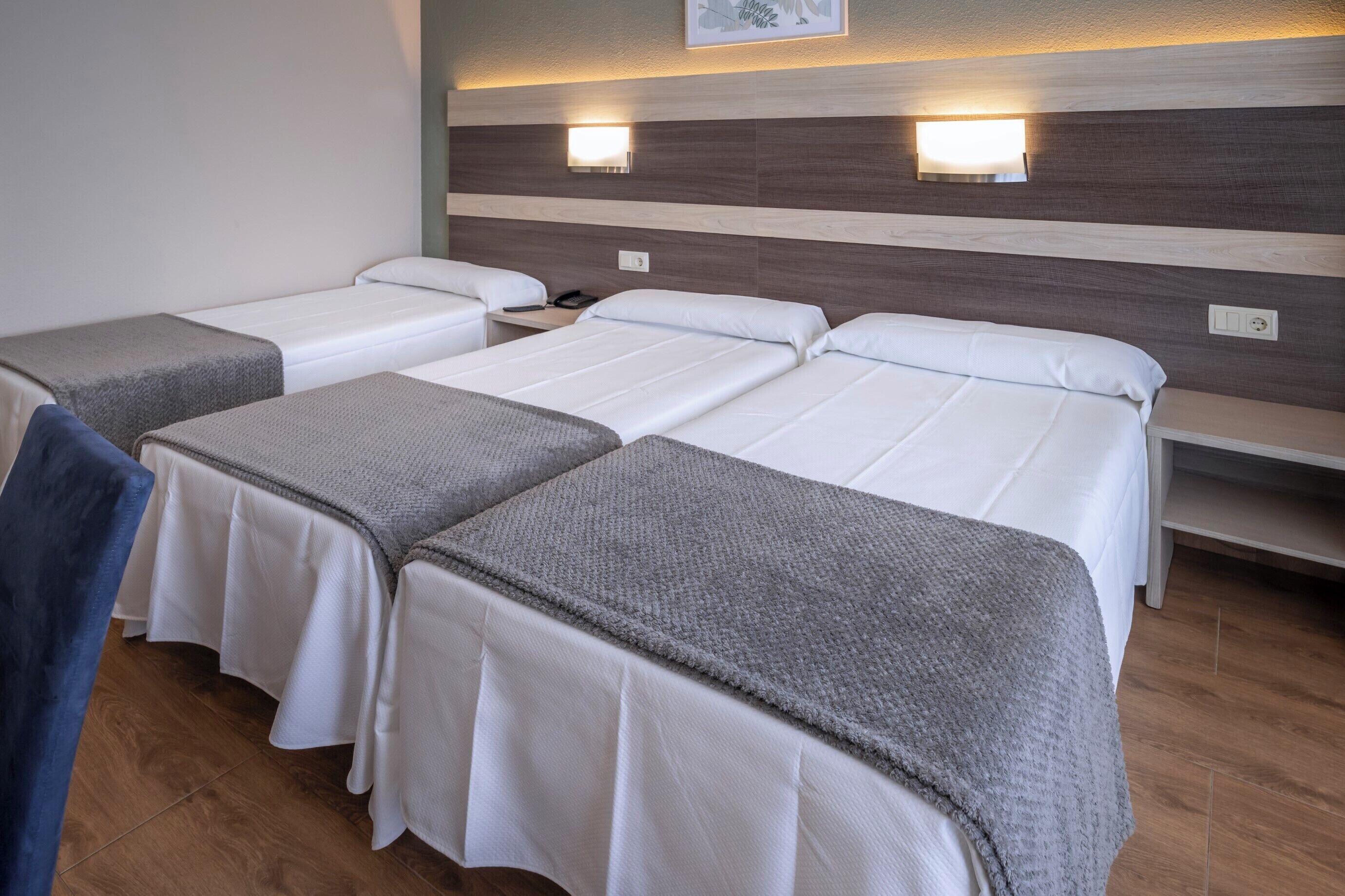 Habitaci ón de hotel con tres camas individuales, cada una con colchón blanco y cabecero gris. Hay luces fijadas a la pared sobre las camas y una pequeña mesa entre ellas.