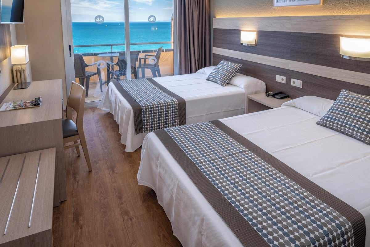 A hotel room with two beds, a desk, and a balcony with a view of the sea.