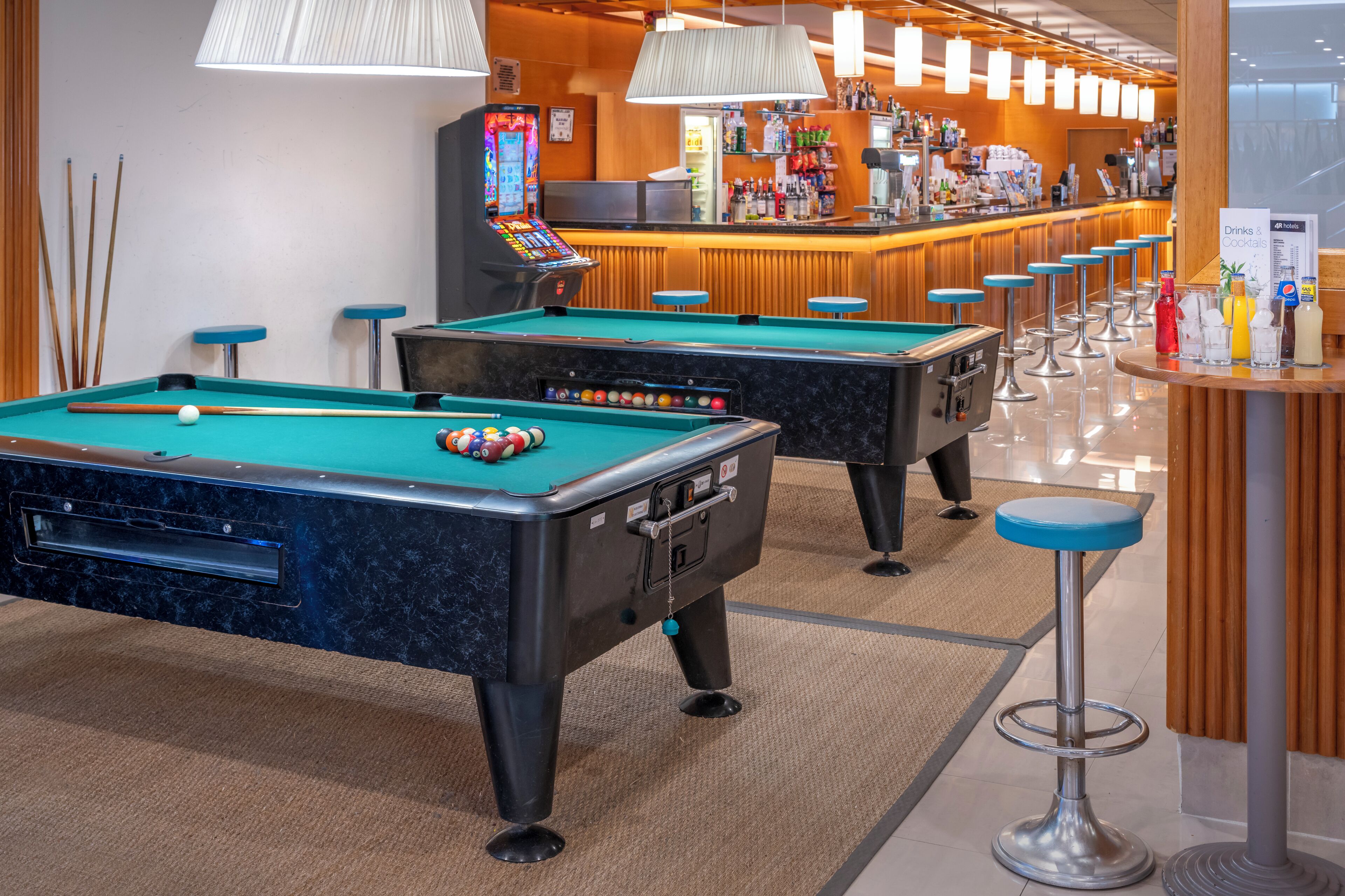 A pool table with a pool cue and balls, a bar area with stools, and a billiard table in a hotel lobby.