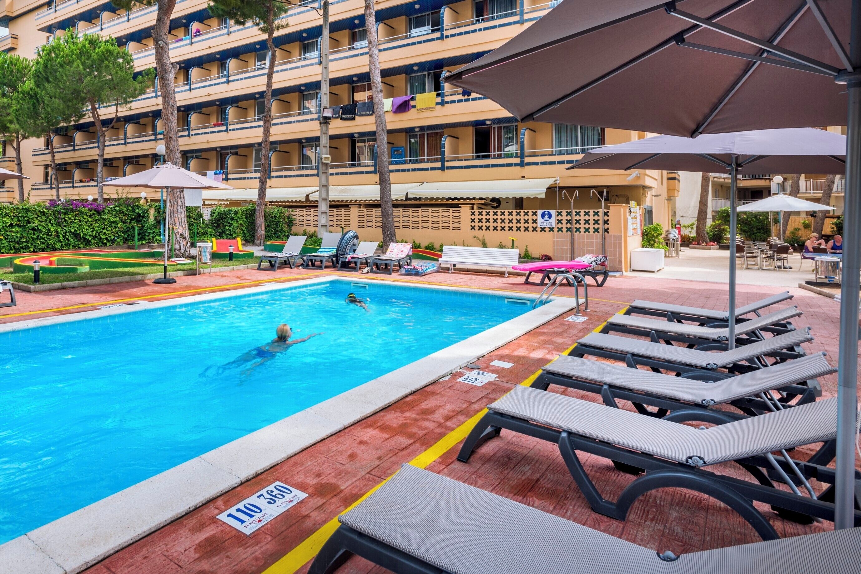 A hotel pool area with lounge chairs, umbrellas, and a person swimming.