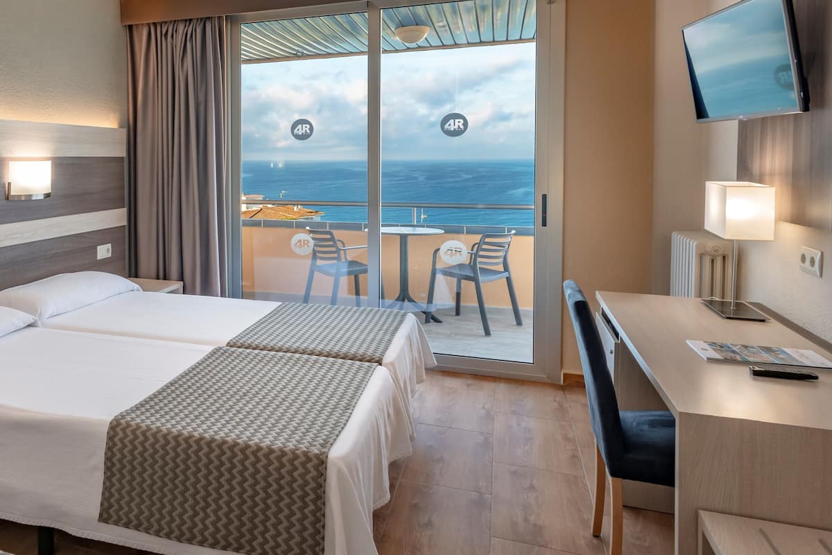 A hotel room with two beds, a desk, a chair, a TV, and a balcony with a view of the sea.