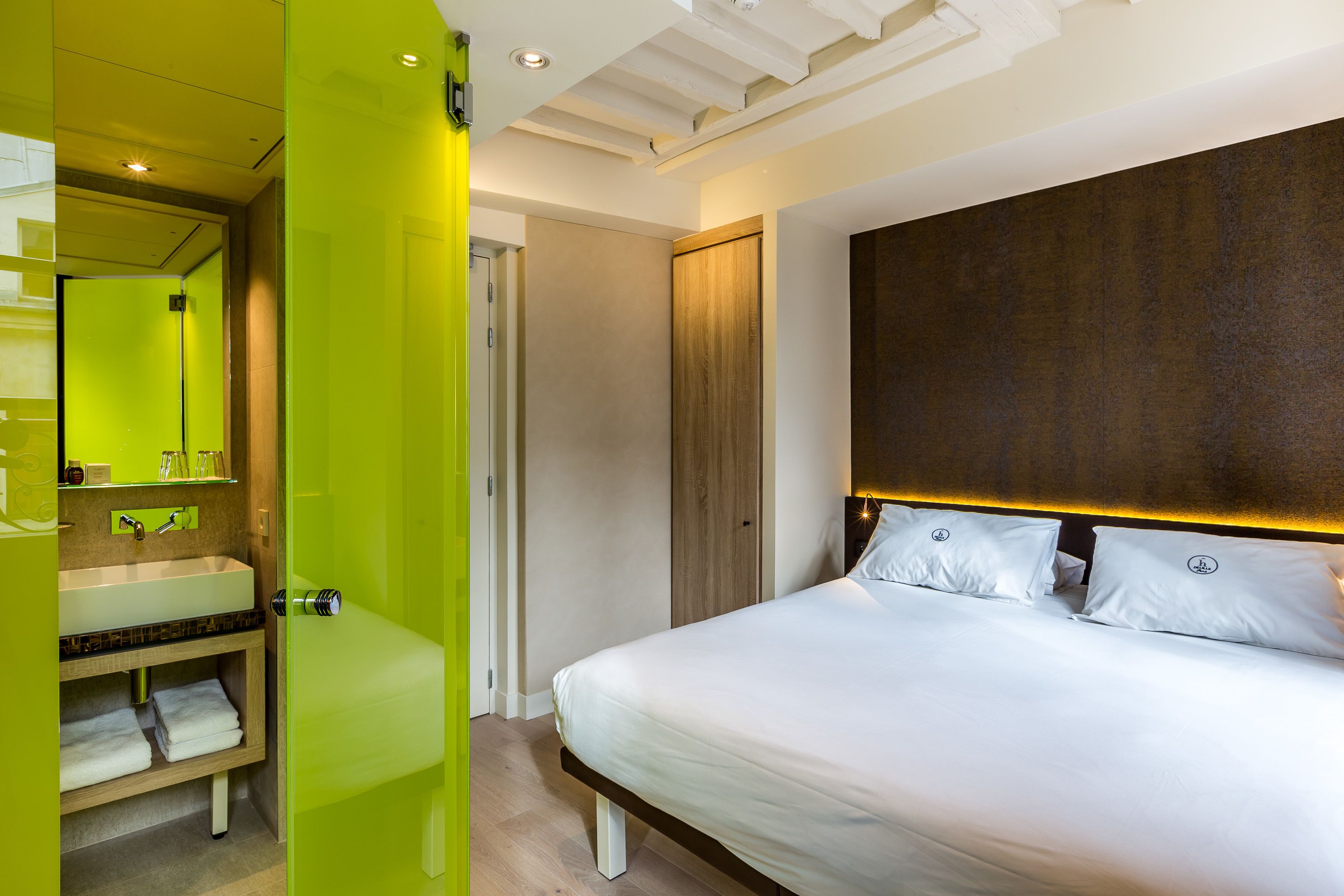 A modern hotel room with a large bed, a bright green wall, a wooden headboard, and a large mirror above the sink.