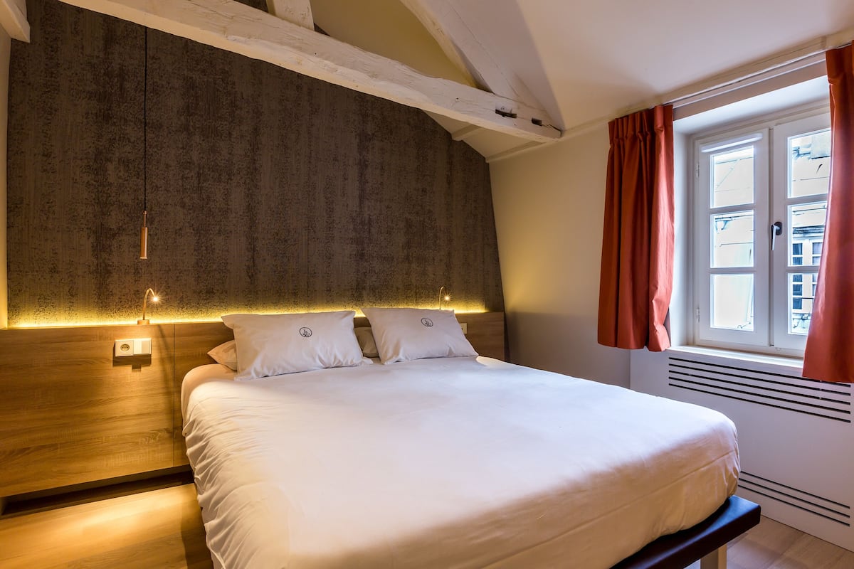 A neatly made bed with a headboard, a bedside table, and a window with curtains.