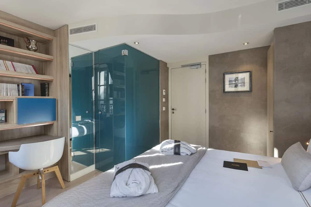 A modern hotel room with a glass shower enclosure, a desk with books, a white chair, and a neatly made bed.