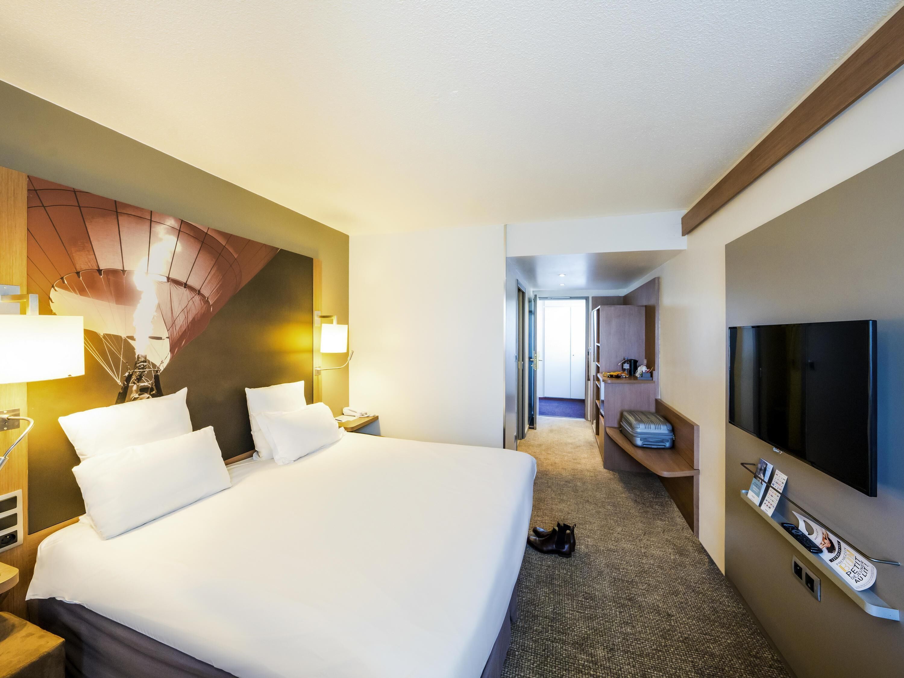 Ein Hotelzimmer mit einem großen Bett, einem Fernseher, einem Schreibtisch mit Computer und einem Kleiderständer.