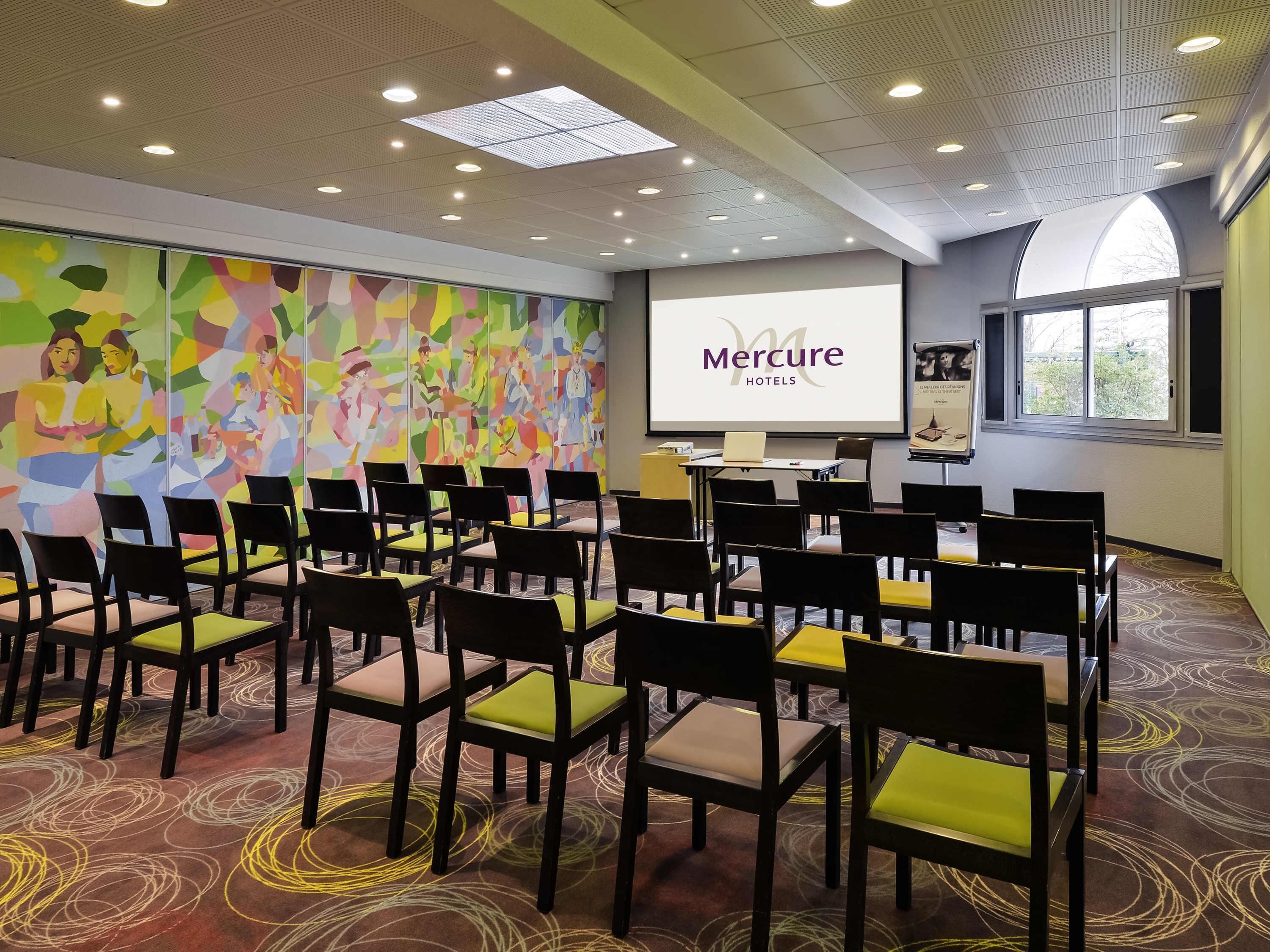 Un salón de conferencias con filas de sillas, una pantalla de presentación y un mural abstracto lleno de color.
