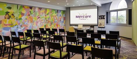 Un salón de conferencias con filas de sillas, una pantalla de presentación y un mural abstracto lleno de color.