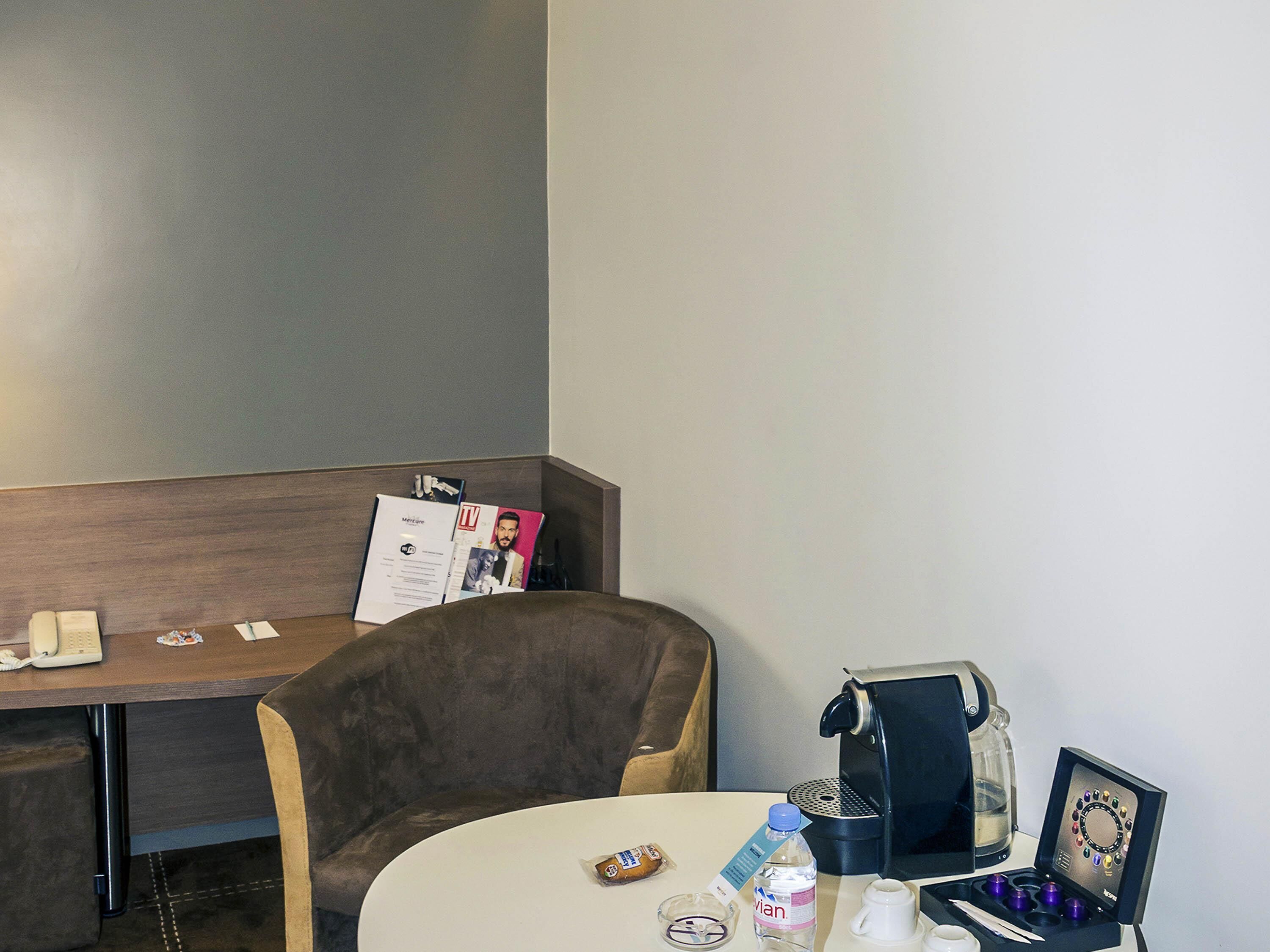 A hotel room with a desk, a chair, a coffee machine, a bottle of water, and a small snack.