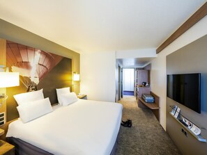 Habitación de hotel con una cama grande, televisión, escritorio con computadora y perchero.
