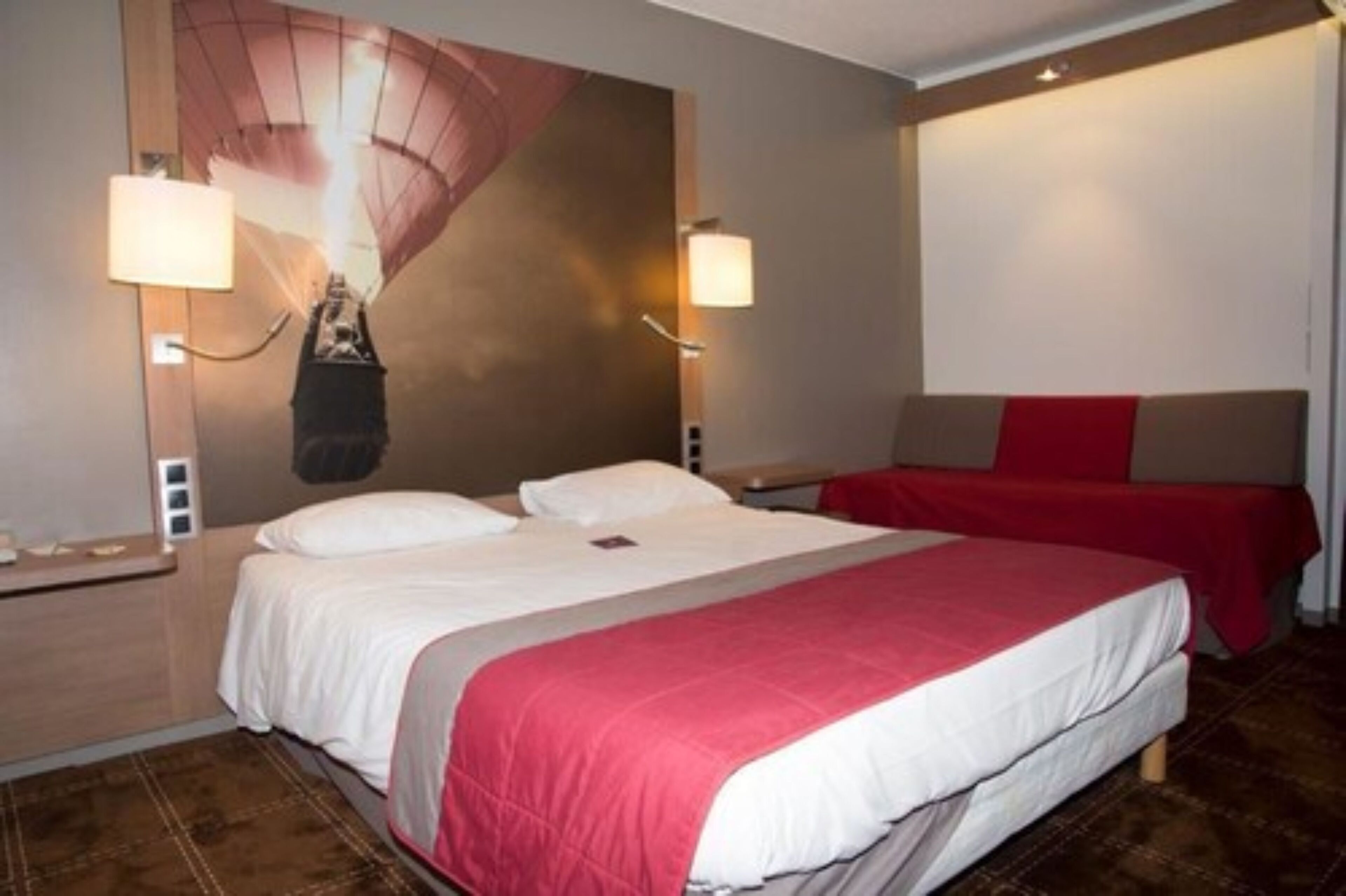 A hotel room with a large bed, two bedside lamps, a red accent wall, and a red throw blanket.