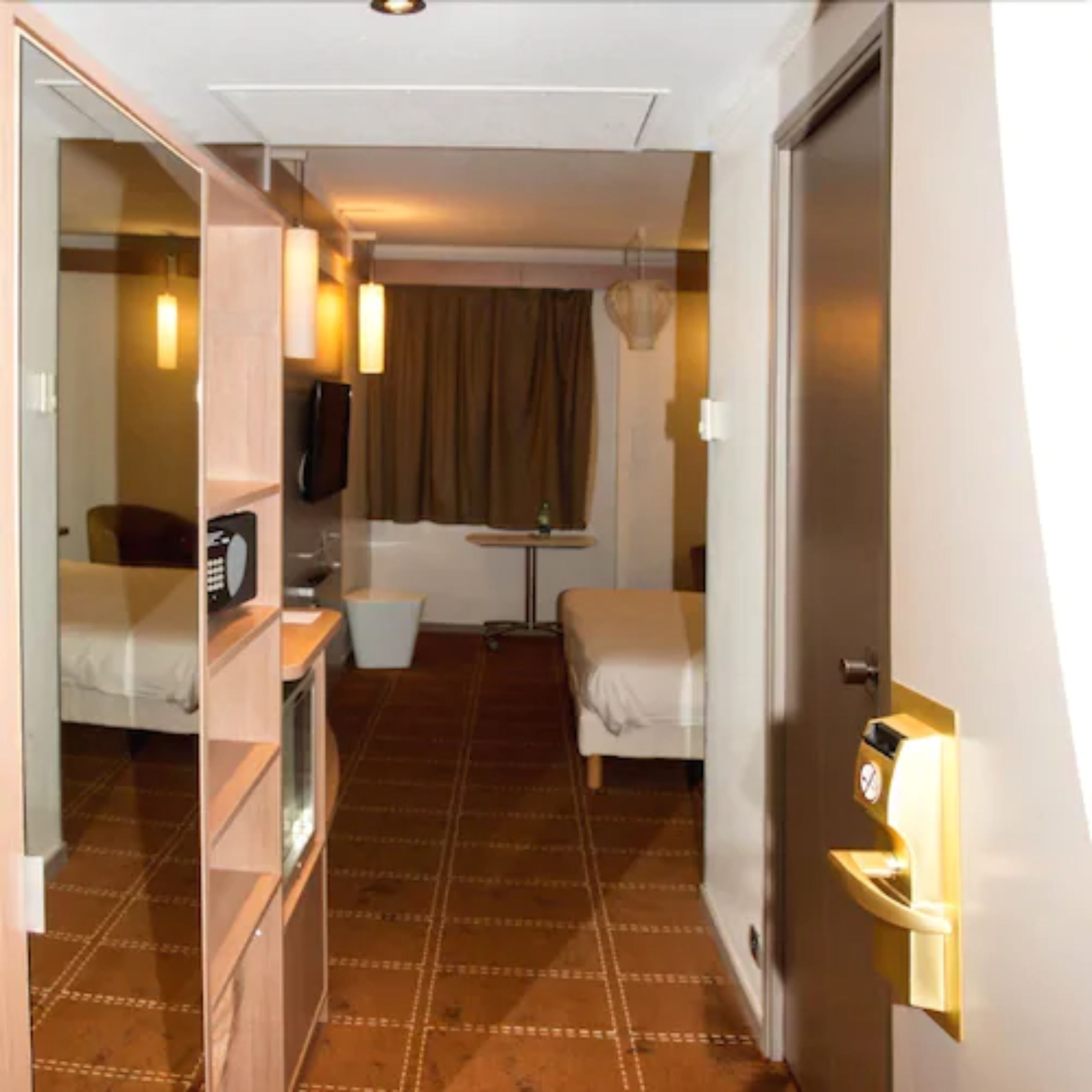 A hotel room with a bed, a desk, a chair, a small table, a mirror, and a door with a light switch.