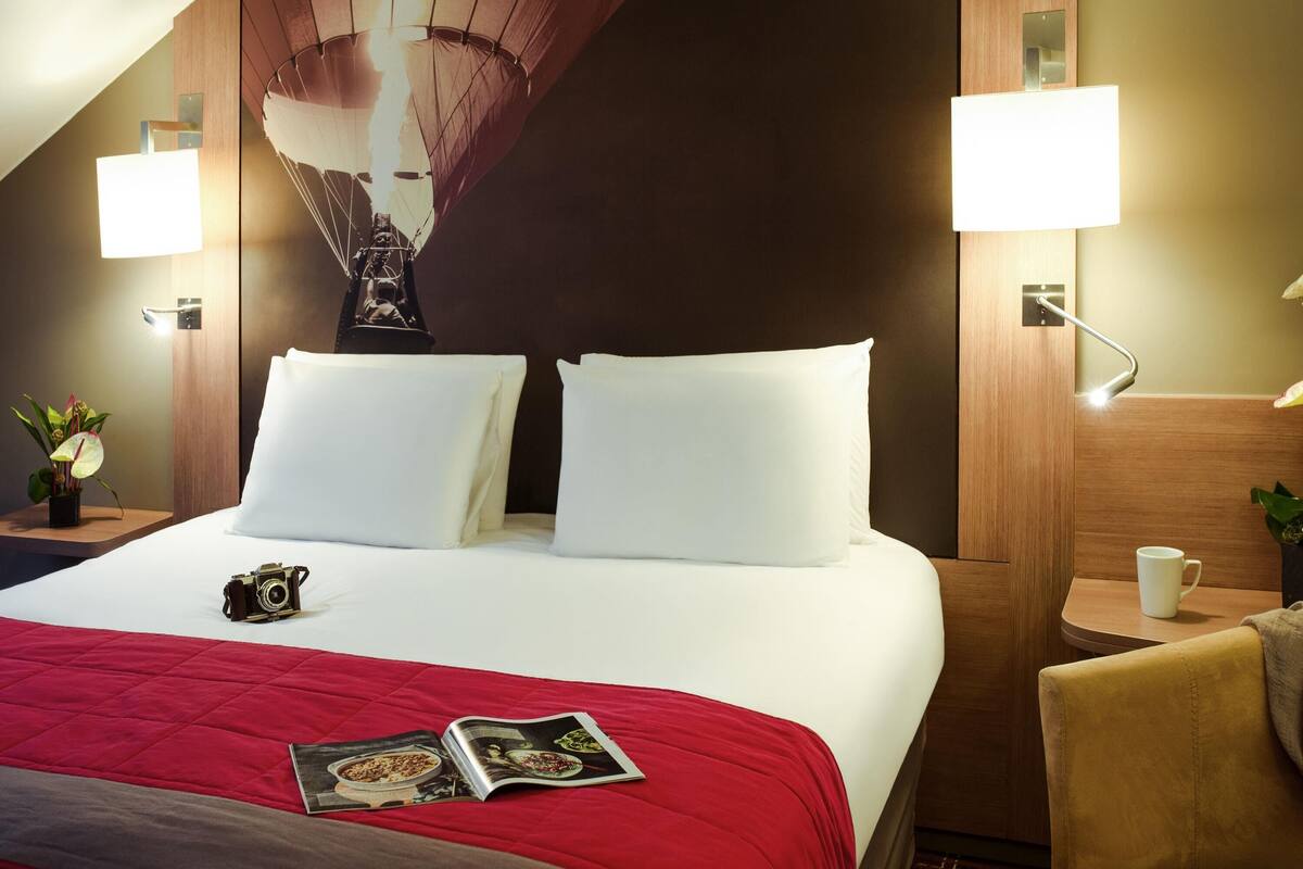 A hotel room with a large bed, two bedside lamps, a nightstand with a lamp, a chair, and a magazine on the bed.