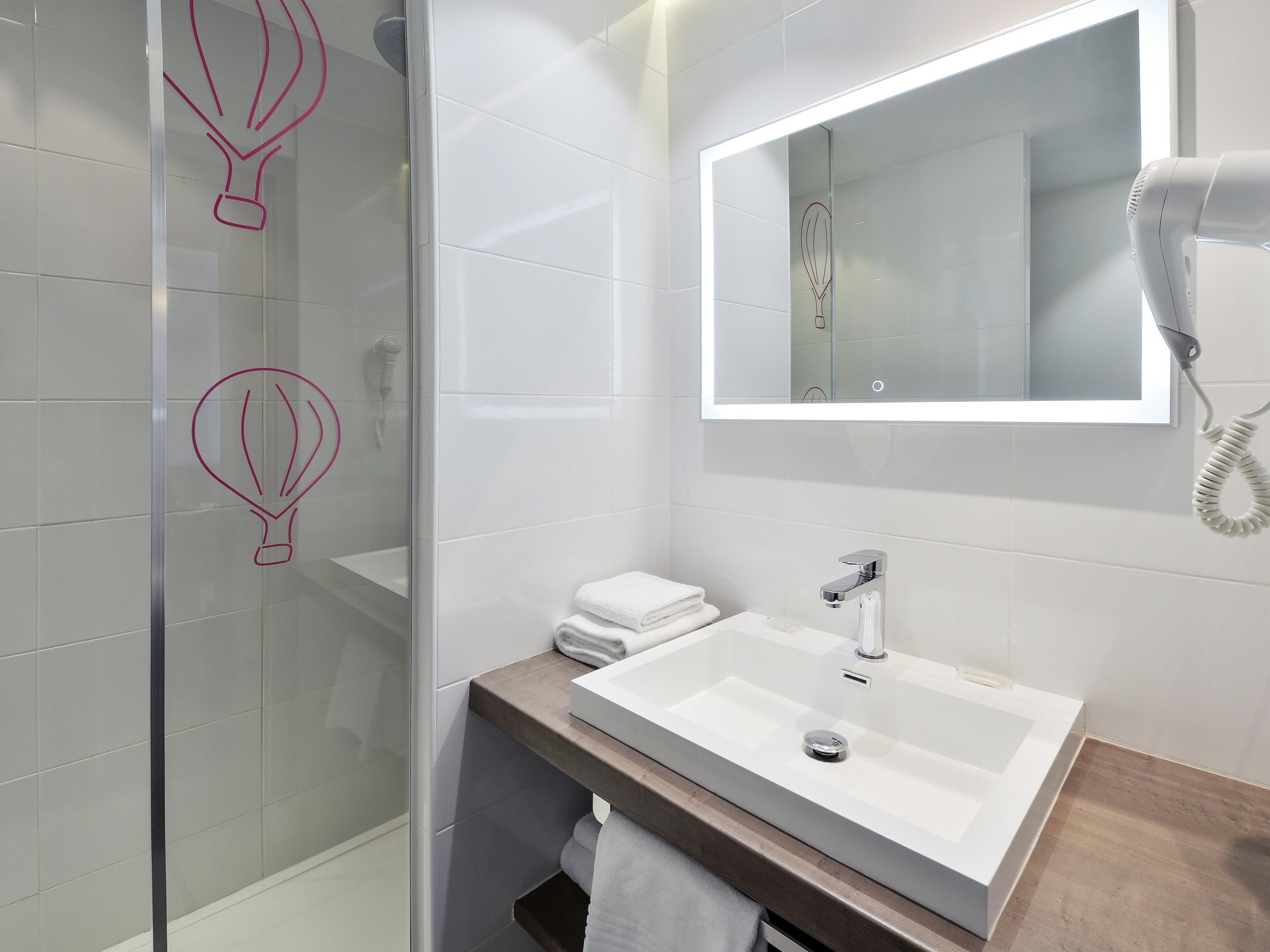 A modern bathroom with a glass shower enclosure, a white sink with a wooden countertop, and a hairdryer mounted on the wall.