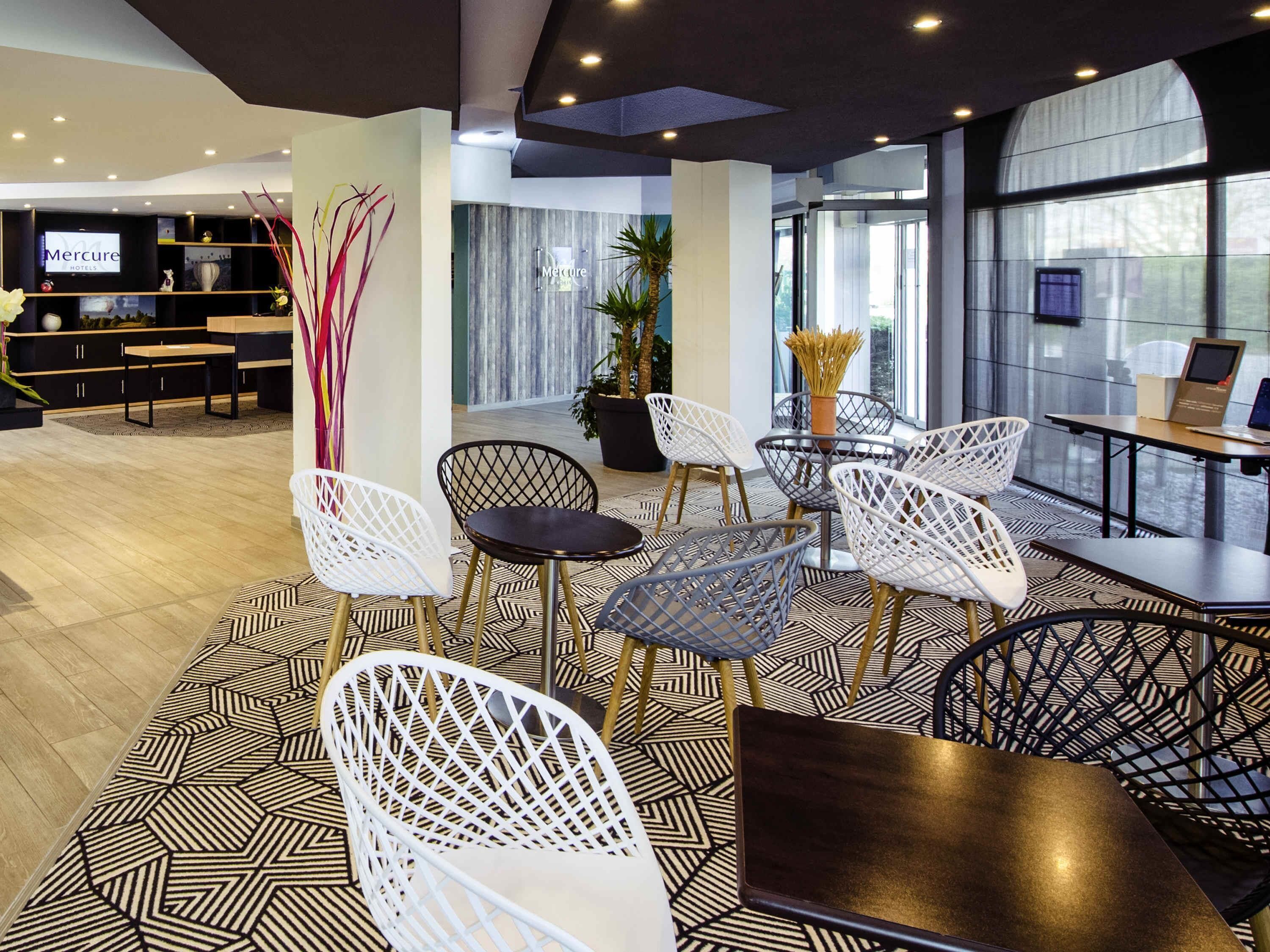 Un hall d’hôtel moderne avec un sol à motifs géométriques, des chaises blanches et noires, un grand écran de télévision et un comptoir d’accueil.