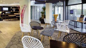 Un hall d’hôtel moderne avec un sol à motifs géométriques, des chaises blanches et noires, un grand écran de télévision et un comptoir d’accueil.