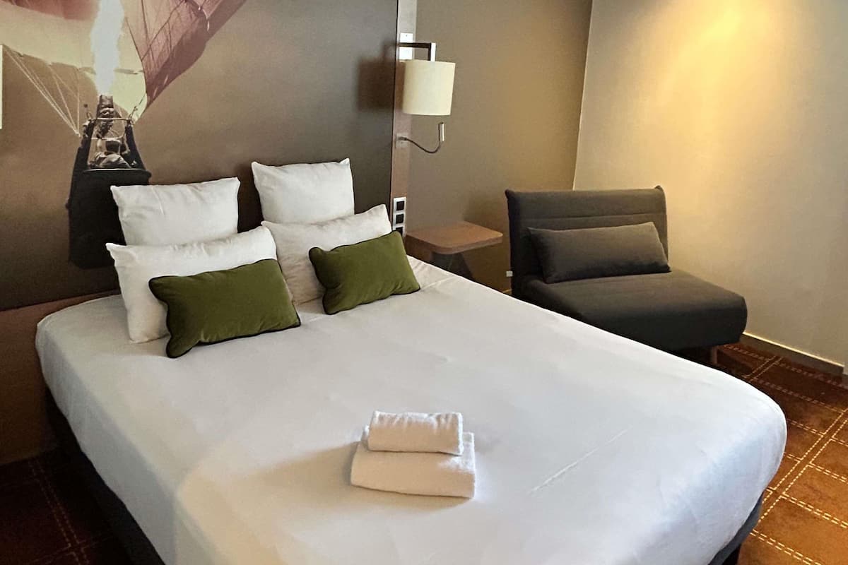 A hotel room with a large bed, a desk, a chair, and a wall-mounted lamp.