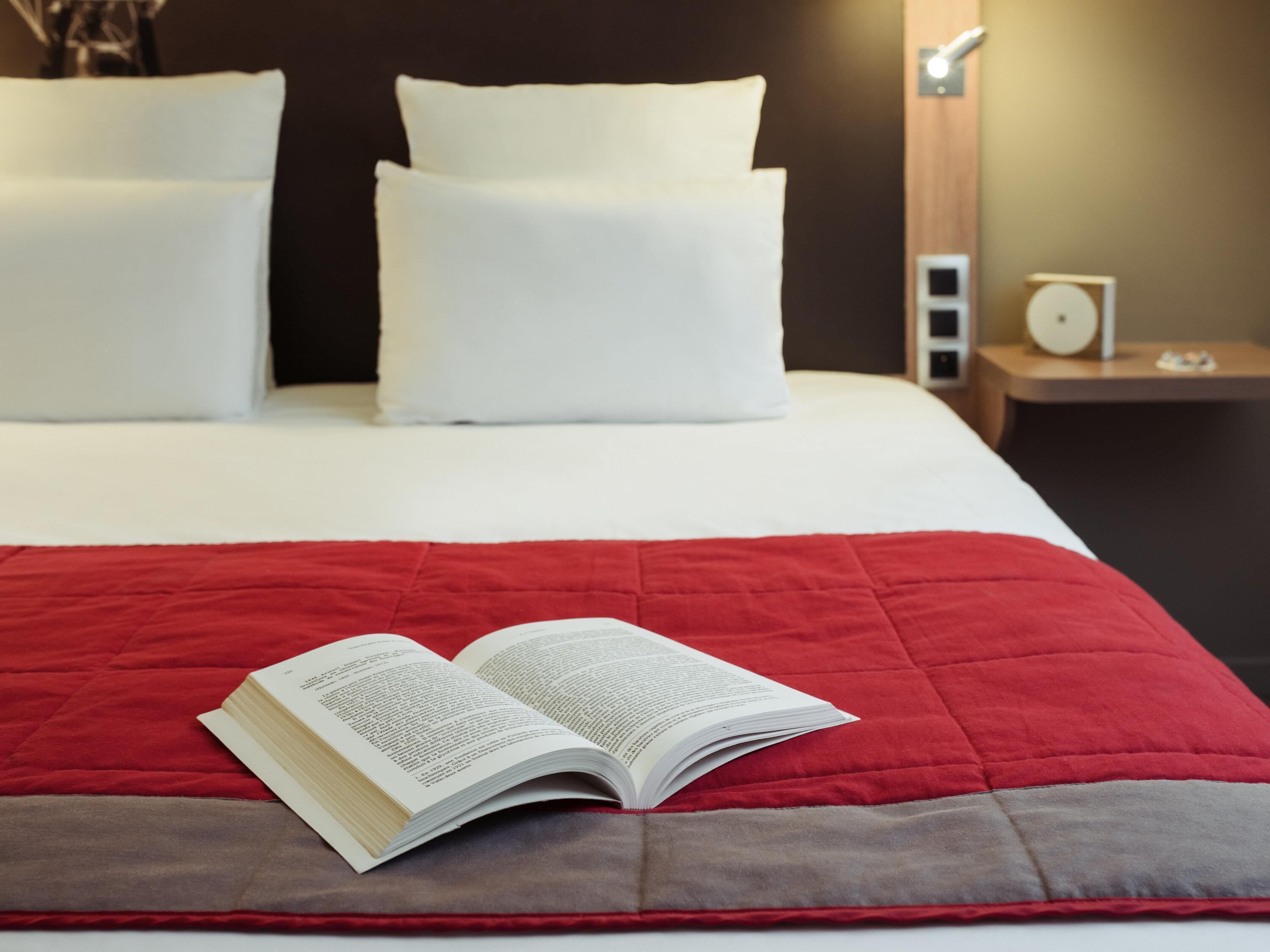 A neatly made bed with a red and gray bedspread, white pillows, and an open book on the bed.