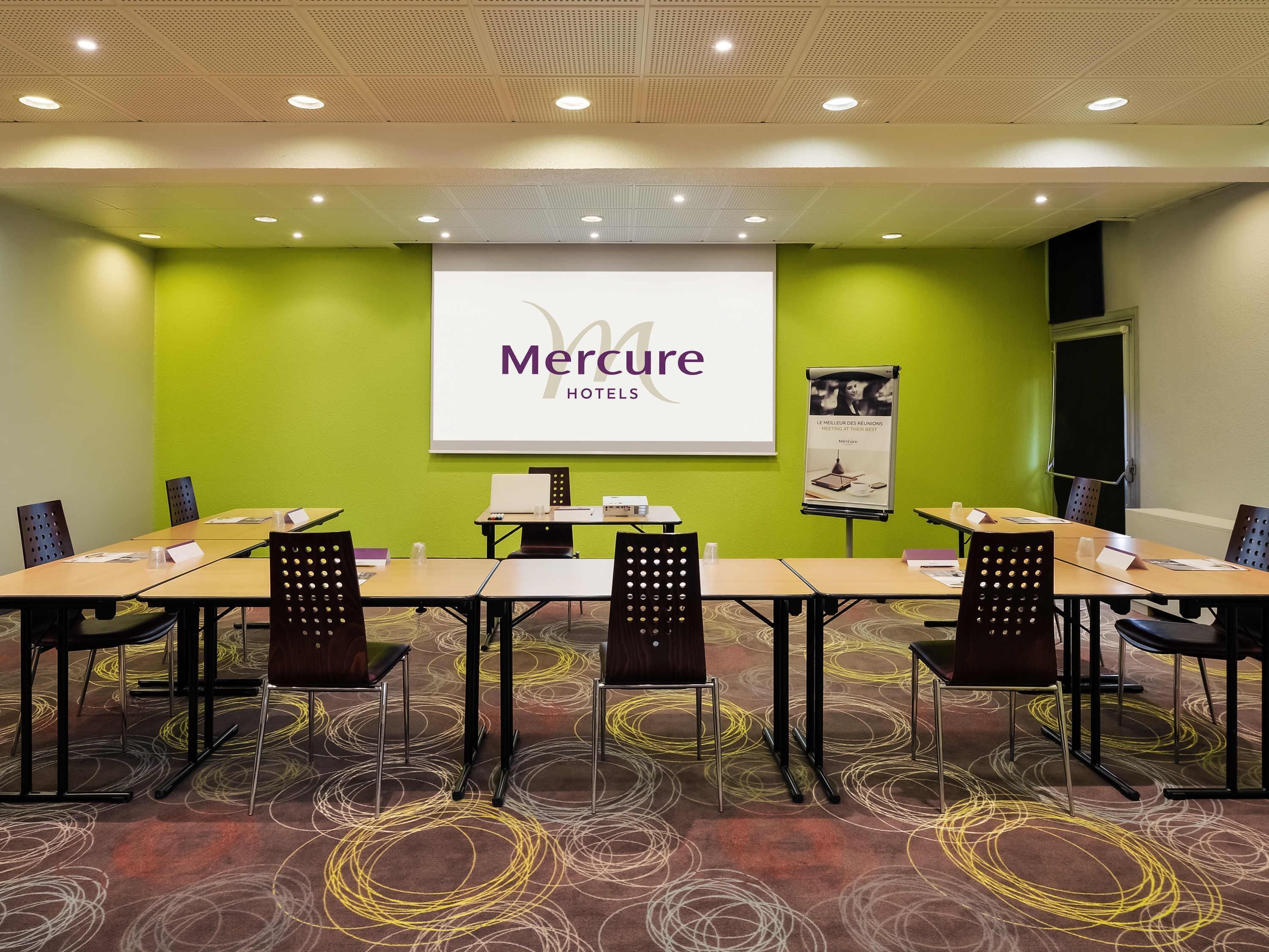 Un salón de conferencias con pared verde, una pantalla de proyector mostrando 'Mercure Hotels' y un cartel.