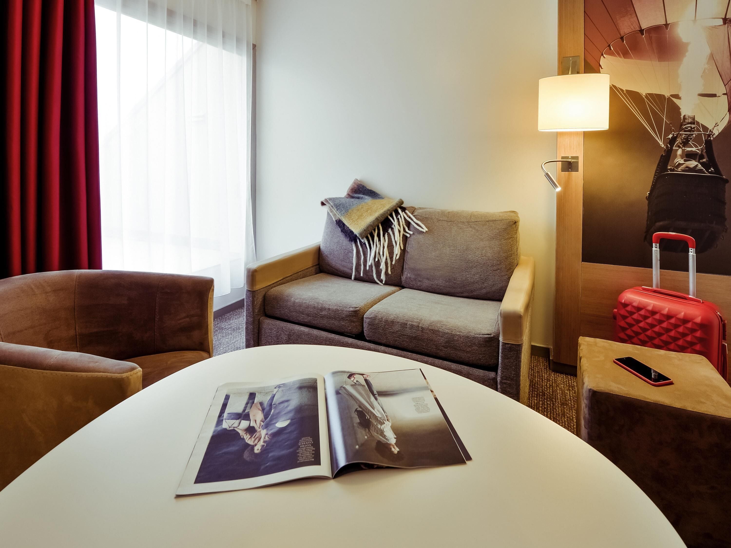 A hotel room with a sofa, armchair, table, magazine, lamp, and a red suitcase.