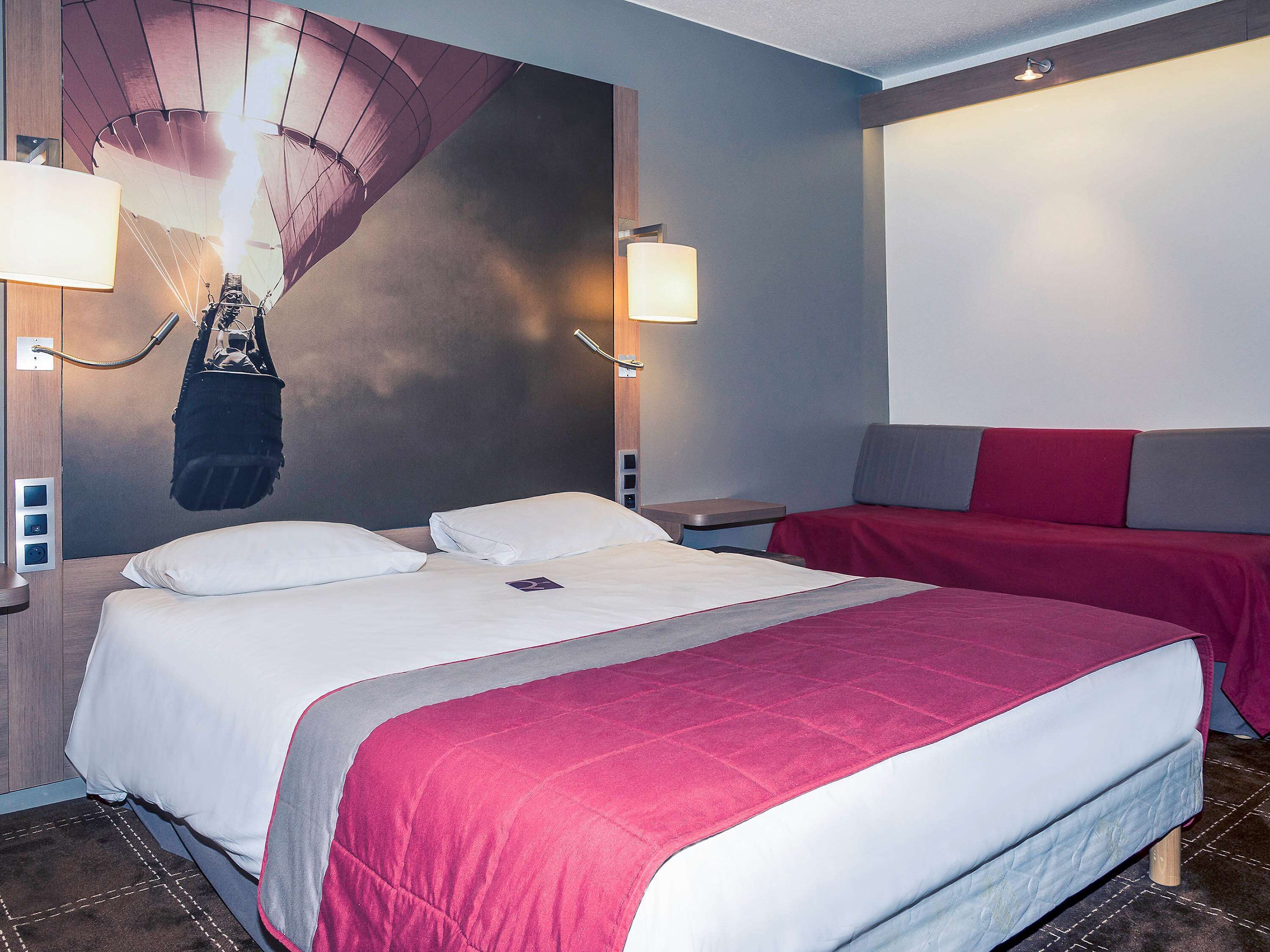 A hotel room with a large bed, two bedside tables, a wall-mounted lamp, and a red and grey sofa.