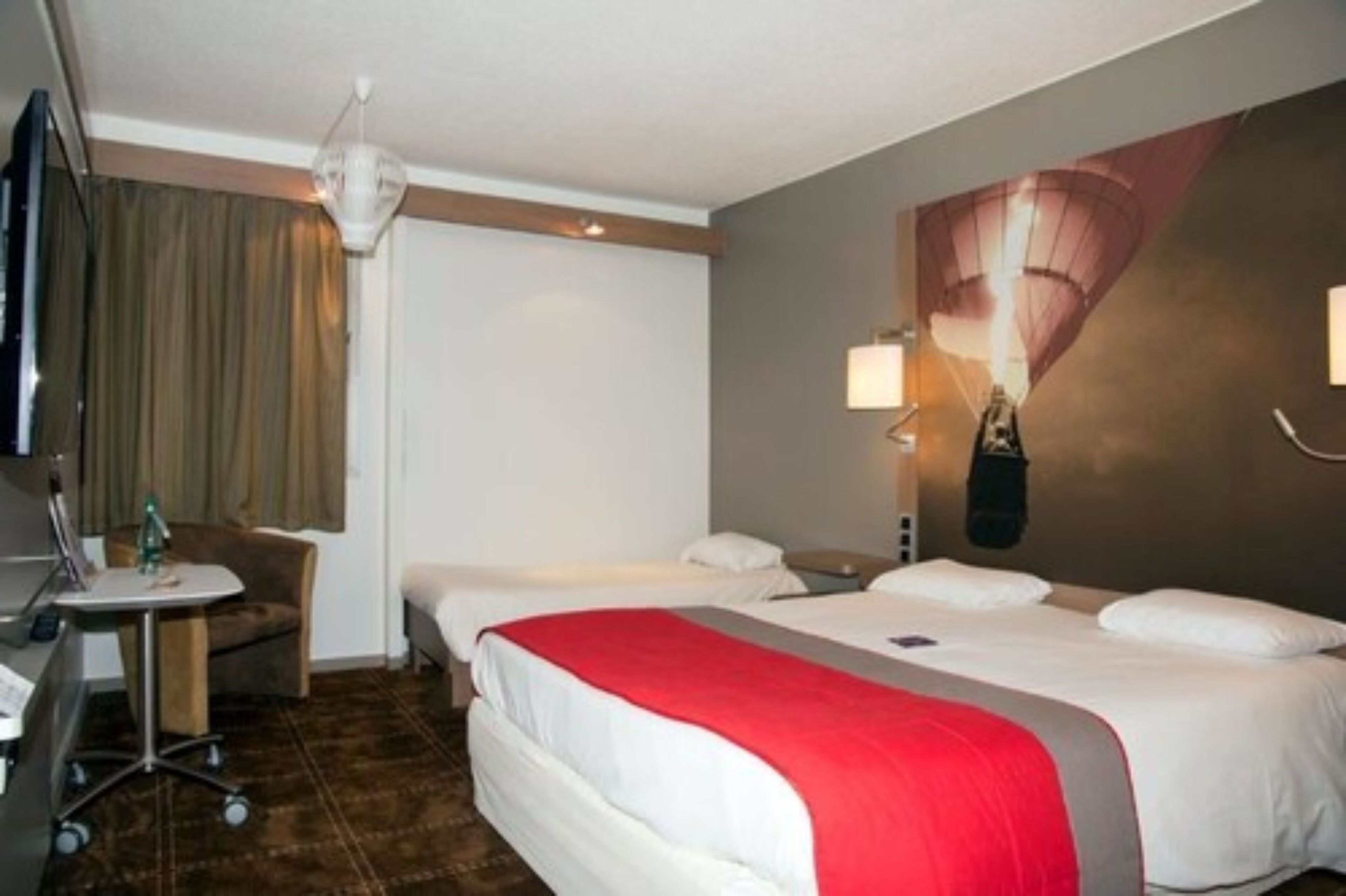 A hotel room with two beds, a desk with a chair, a television, and a wall-mounted lamp.