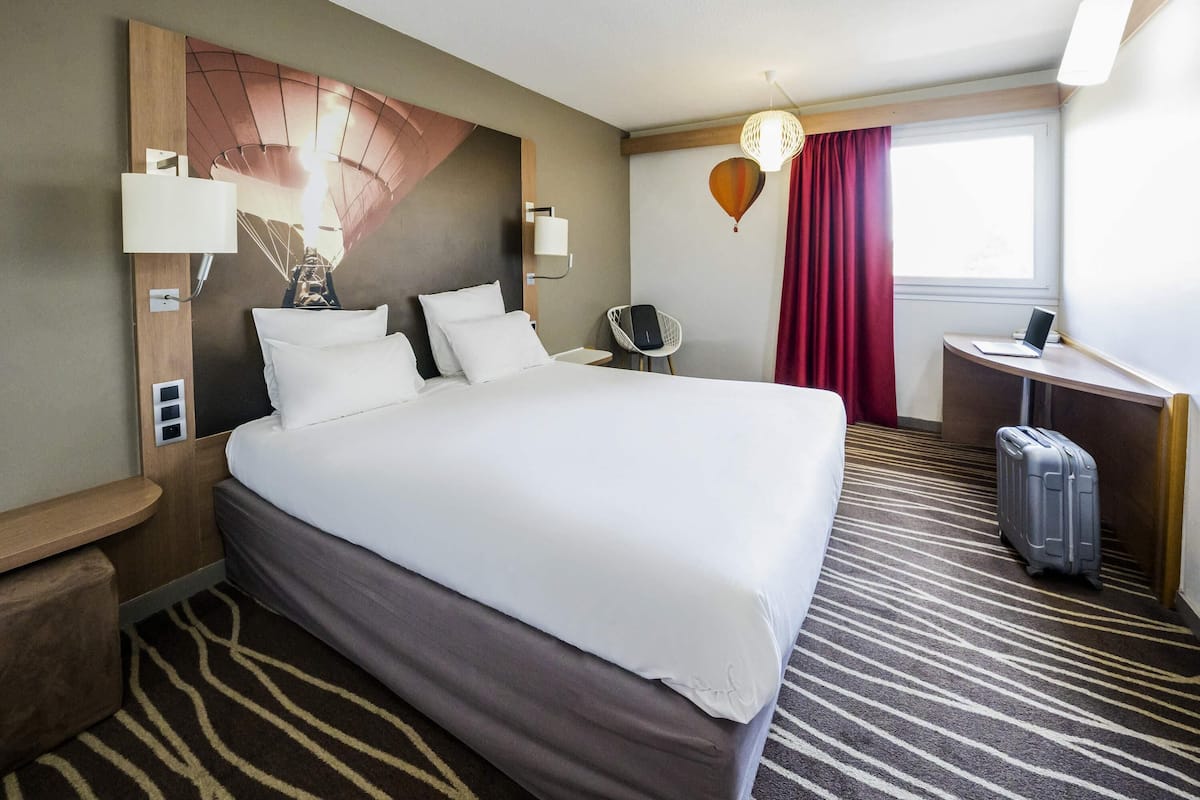 A hotel room with a large bed, a desk, a chair, a lamp, and a suitcase.