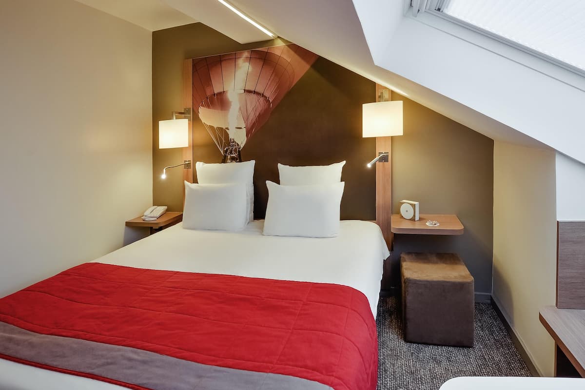 A hotel room with a bed, two bedside tables, a nightstand, a red bedspread, and a large window with blinds.