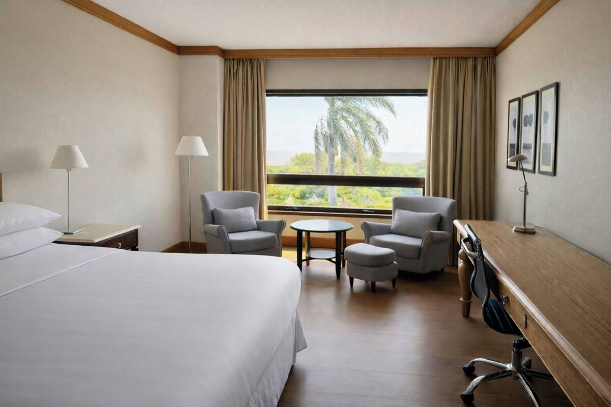 Habitación de hotel con cama, dos sillones, una mesita redonda, un escritorio y vistas a palmeras a través del ventanal.