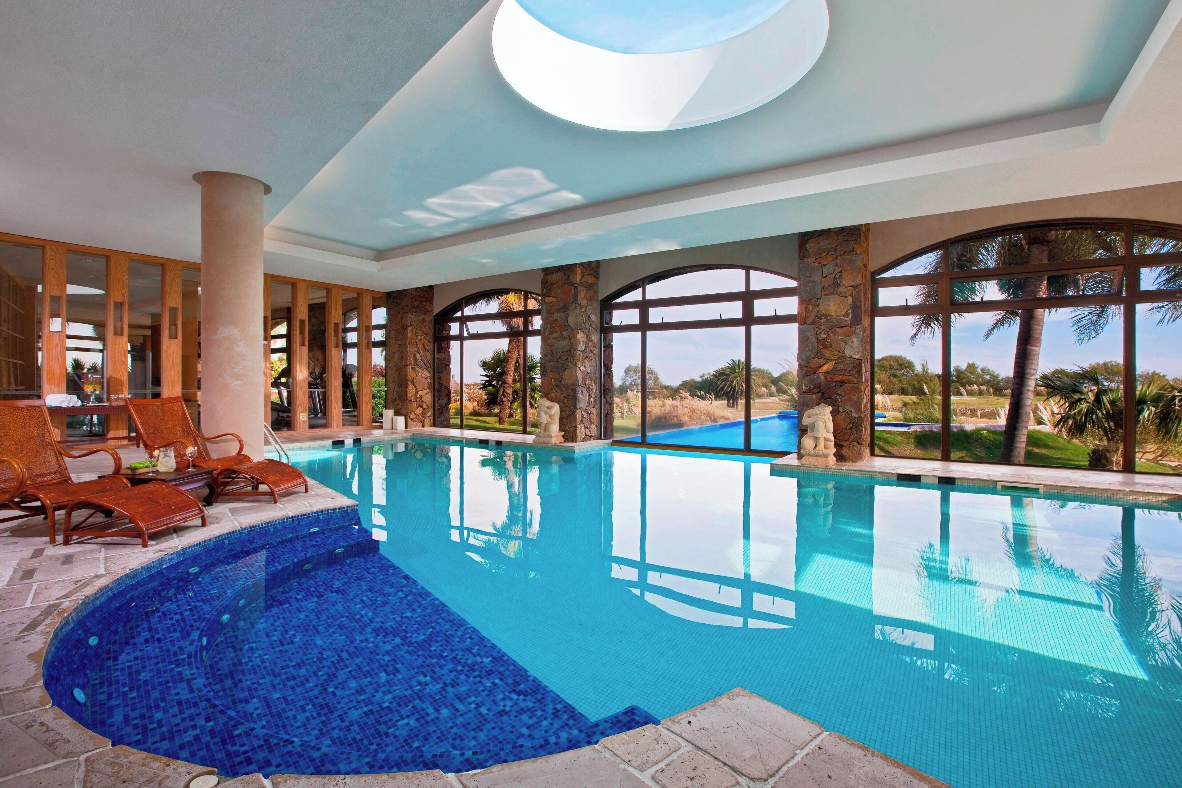 Una piscina interior con paredes de vidrio transparente, pilares de piedra y sillones de descanso.