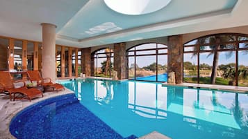Una piscina interior con paredes de vidrio transparente, pilares de piedra y sillones de descanso.