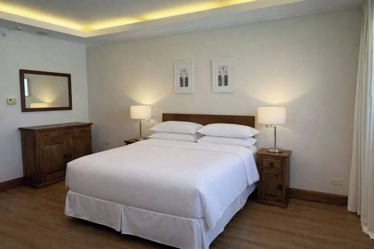 Habitación de hotel ordenada con una cama grande, dos mesitas de noche con lámparas, un aparador de madera y cuadros enmarcados en la pared.