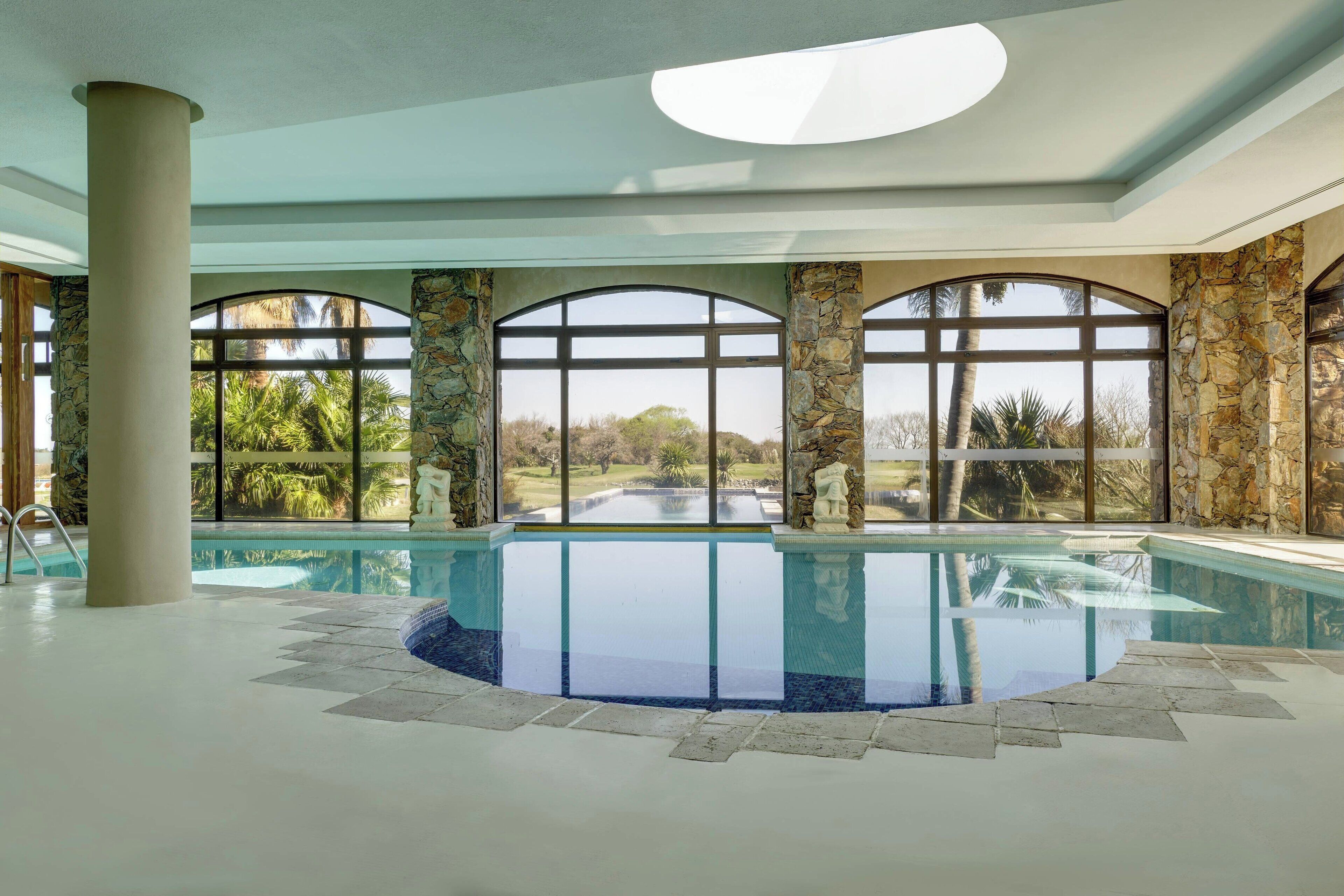 Una piscina interior con columnas de piedra y amplios ventanales de arco que dan a un jardín.