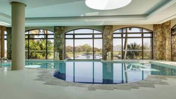 Una piscina interior con columnas de piedra y amplios ventanales de arco que dan a un jardín.