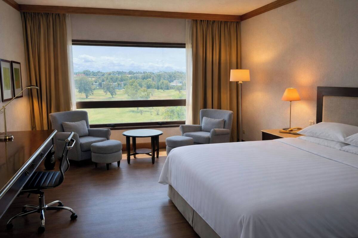 Habitación de hotel con cama, dos sillones, escritorio, lámpara y vistas a un paisaje verde.