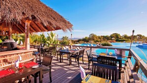 A wooden deck with tables and chairs, a thatched-roof structure, and a pool with lounge chairs.