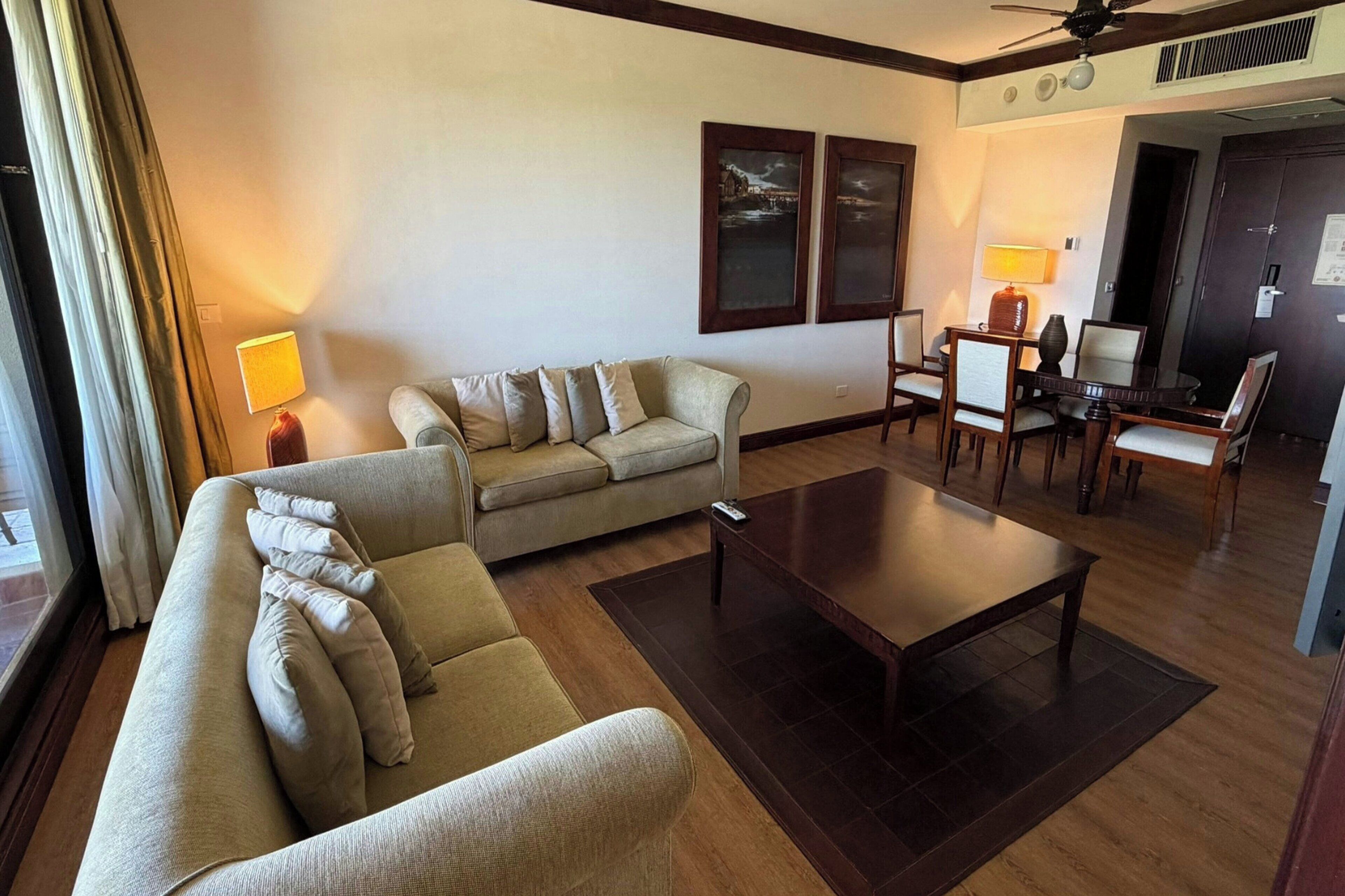 A hotel living room with a sofa, a coffee table, and a dining area with chairs and a table.